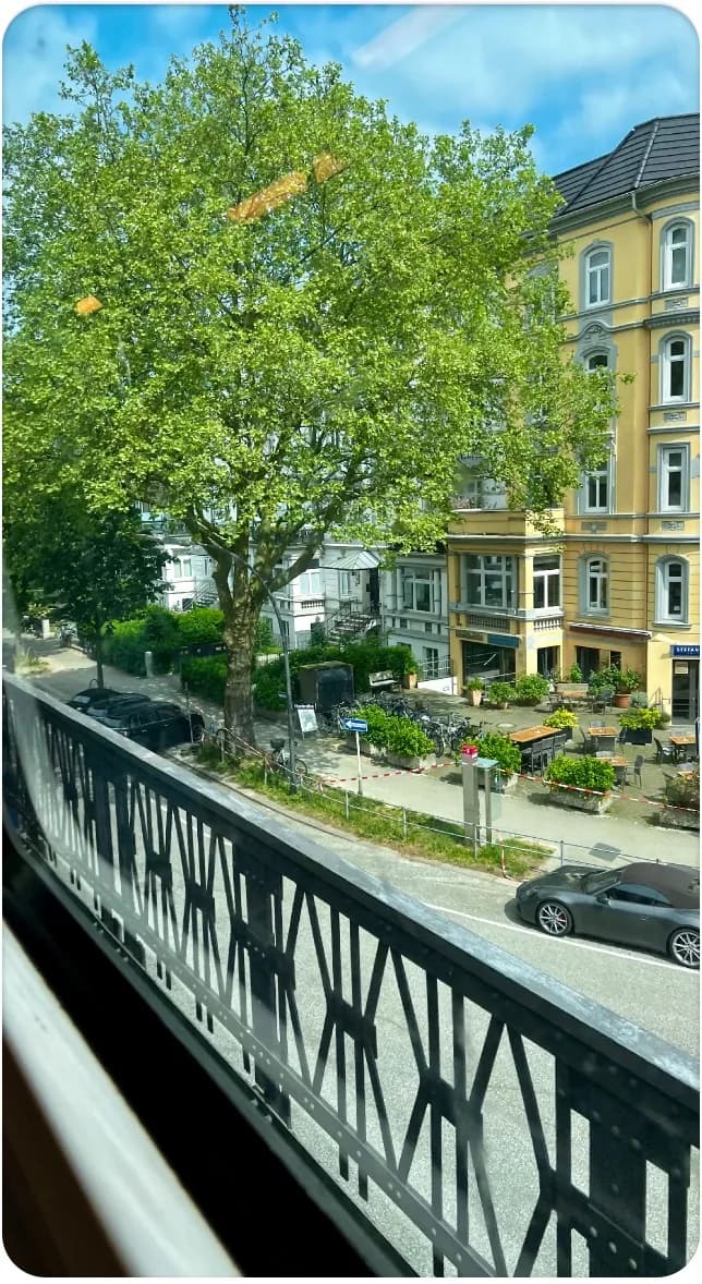 A leafy Hamburg street with a yellow apartment building and a pavement cafe, seen from a train window over an iron bridge railing.