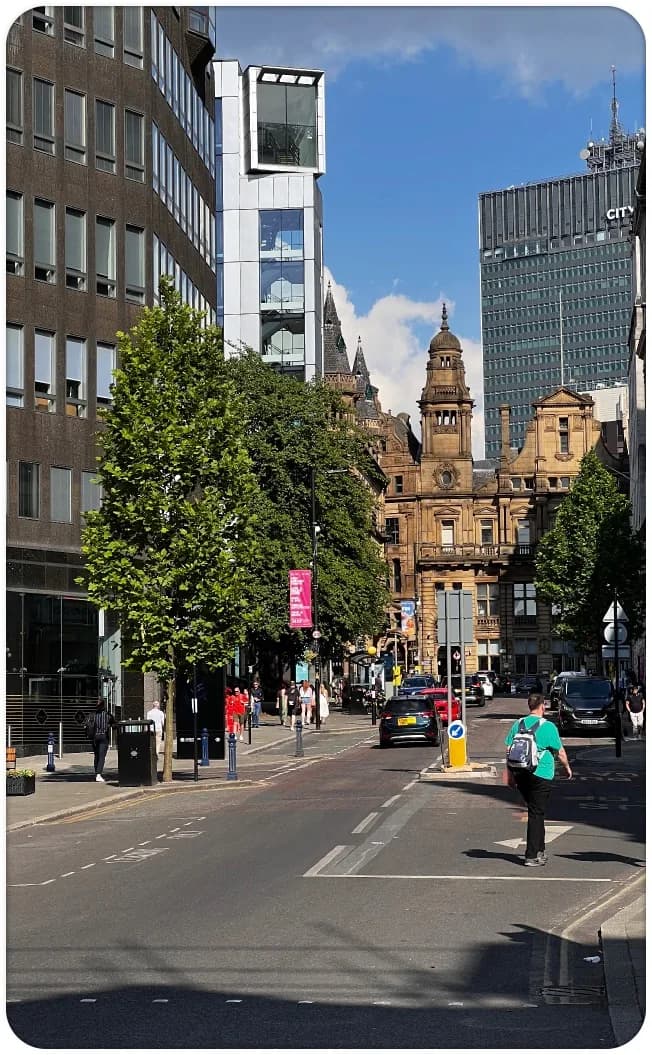 A sunny Manchester street looking toward Victorian sandstone buildings and a glass tower, with a man crossing the road.